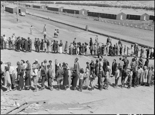 San Bruno, California. This assembly center has been open for two days. Bus-load after bus-load of evacuated persons of Japanese ancestry are arriving on this day after going through the necessary procedures, they are guided to the quarters assigned to them in the barracks. Only one mess hall was operating today. Photograph shows line-up of newly arrived evacuees outside this mess hall at noon. Note barracks in background, just built, for family units. There are three types of quarters in the center of post office. The wide road which runs diagonally across the photograph is the former racetrack.